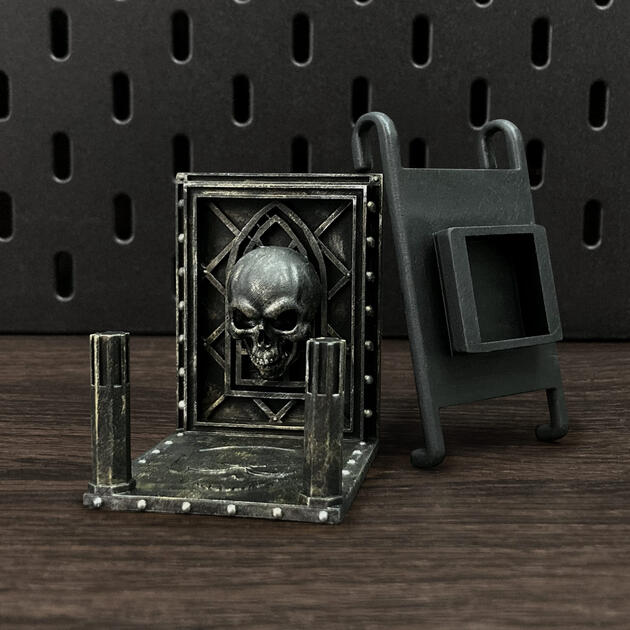 Hand-painted weathered iron skull dice box, mounted on an IKEA SKÅDIS pegboard for TTRPG miniature display.