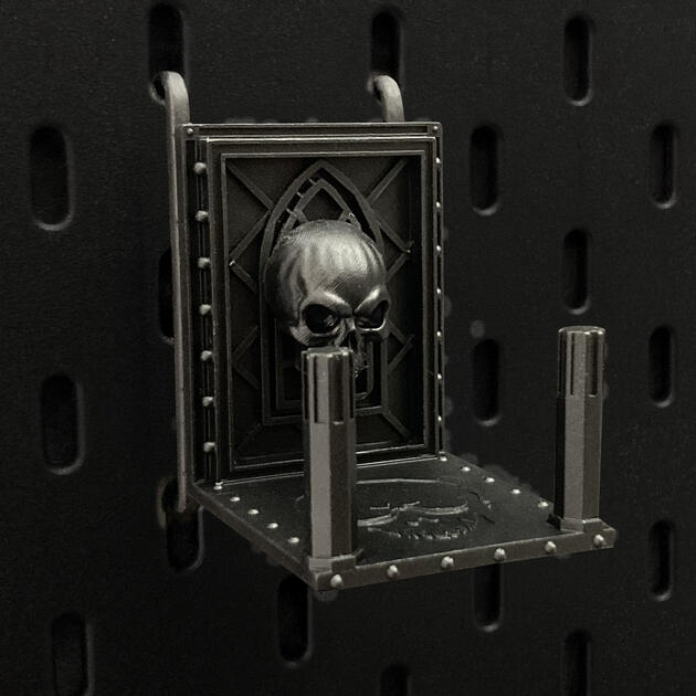Hand-painted weathered iron skull dice box, mounted on an IKEA SKÅDIS pegboard for TTRPG miniature display.