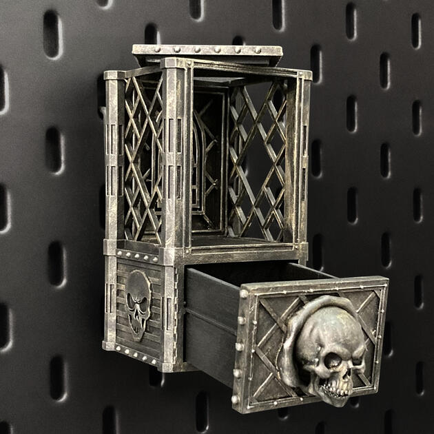Hand-painted weathered iron skull dice box with open storage drawer, mounted on an IKEA SKÅDIS pegboard for TTRPG miniature display.