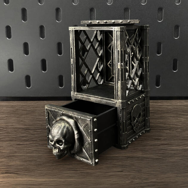 Hand-painted weathered iron skull dice box with open storage drawer, mounted on an IKEA SKÅDIS pegboard for TTRPG miniature display.